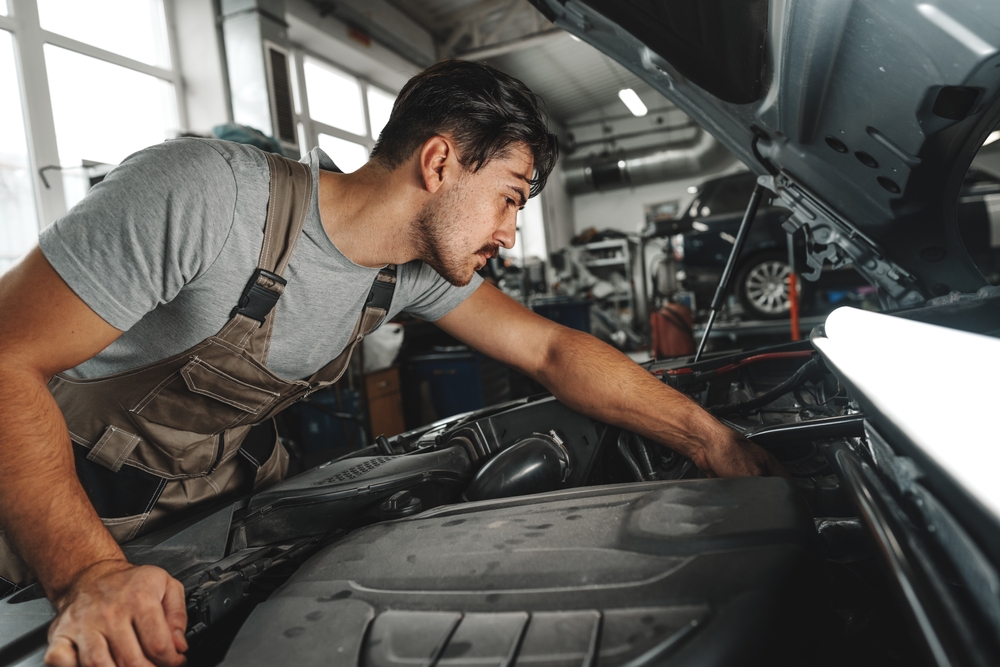 Young,Male,Mechanic,Examining,Engine,Under,Hood,Of,Car,At